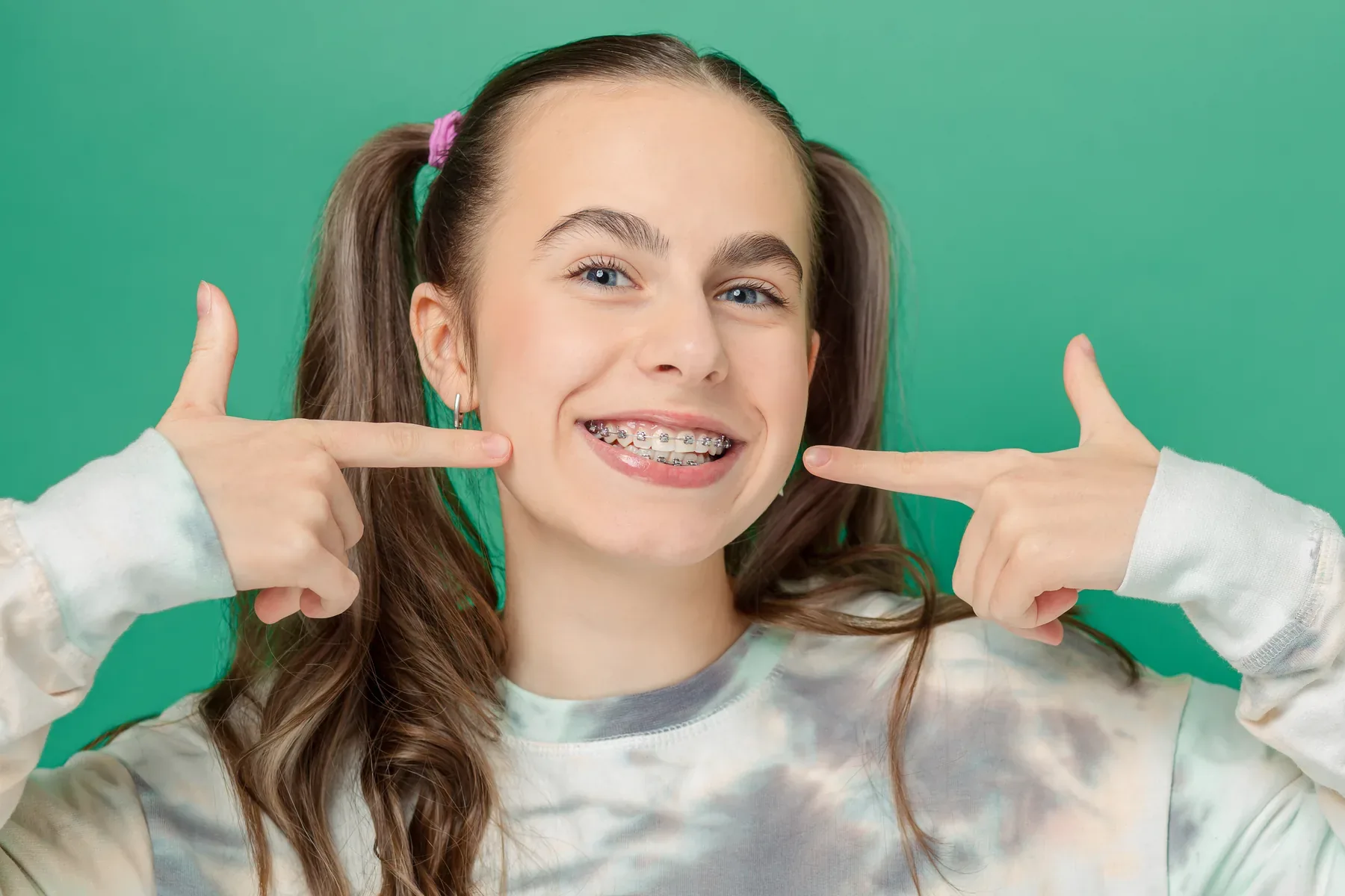 A smiling teen with braces pointing to her teeth